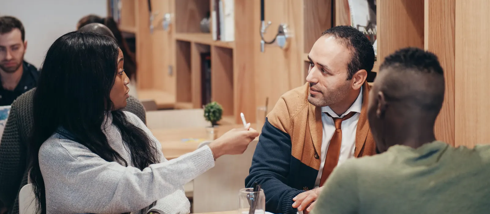 Four people talking around a table in a wood-paneled workspace, one gesturing with a pen while the others listen